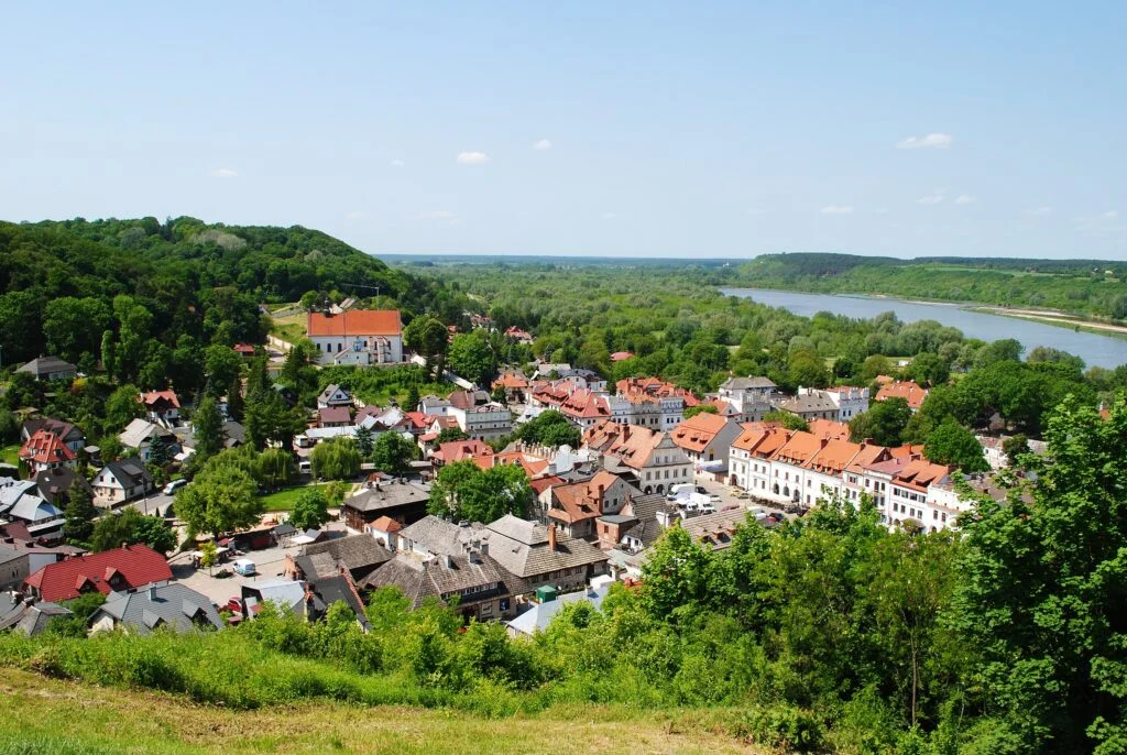 Kazimierz Dolny Poland travel guide – panoramic view of the Renaissance town square with pastel facades, historic church spires, and wooded hills rising beyond the Vistula River under a soft morning sky