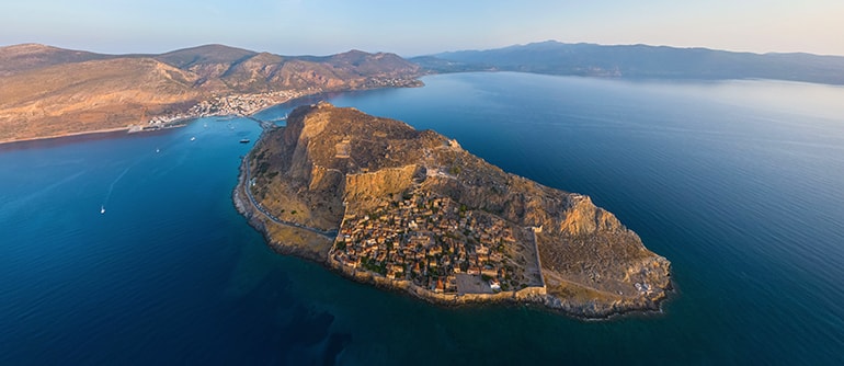 A panoramic view of Monemvasia, Greece with its medieval stone walls, terracotta rooftops, and the shimmering Aegean Sea—ideal for a Monemvasia Greece travel guide.